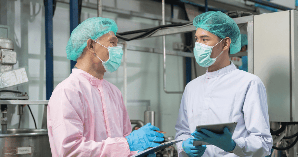 Two capsule filling machine operators wearing PPE, holding clipboards, and performing quality assurance testing.